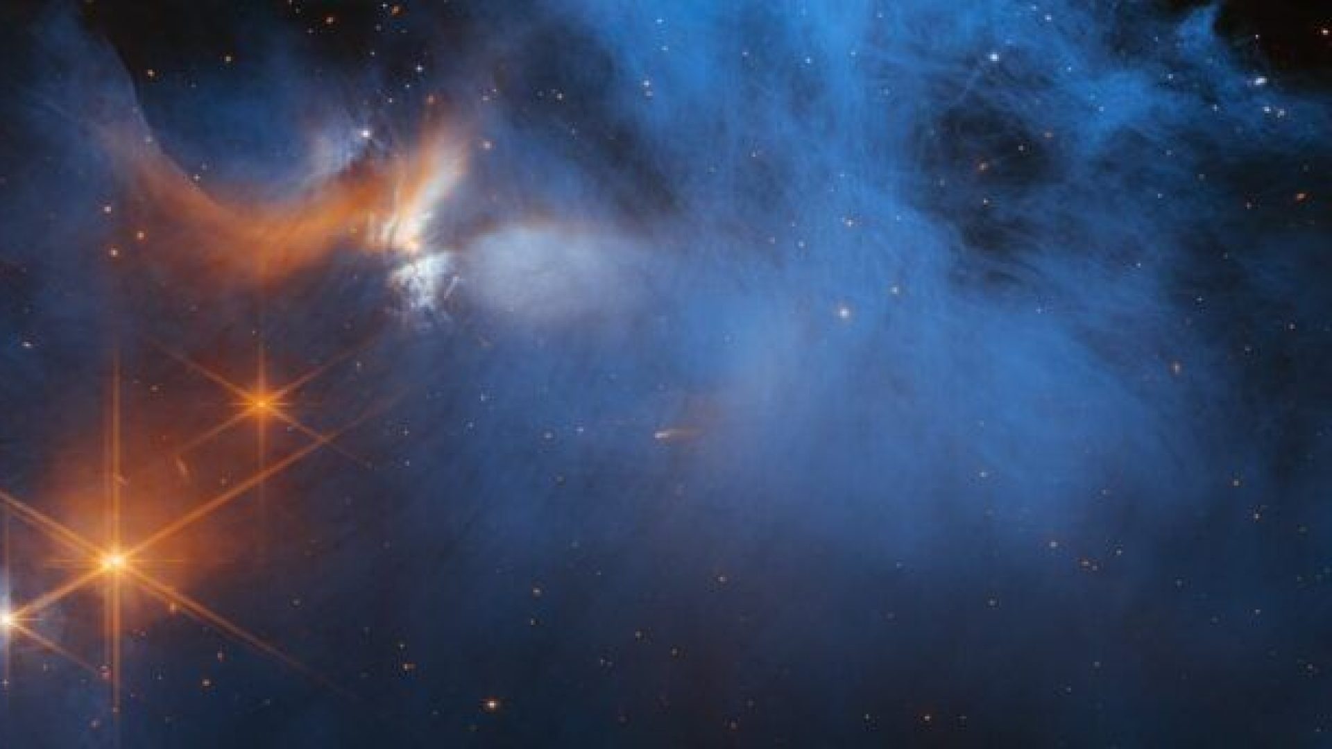 Una grande nuvola scura la cui metà superiore è strutturata come fumo e presenta sottili fessure. Mentre la parte inferiore e ai lati svanisce gradualmente alla vista. Sulla sinistra ci sono diverse stelle arancioni, ciascuna con sei grandi punte, e una dietro la nuvola che la colora di blu pallido e arancione. Sono visibili molte minuscole stelle e lo sfondo è nero. Ph. Credit: NASA, ESA, CSA e M. Zamani (ESA/Webb); Scienze: F. Sun (Steward Observatory), Z. Smith (Open University) e il team ERS dell'era glaciale.